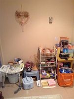 Photo of various bathroom and personal care items arranged against a wall, including plastic stools, handheld massager, basket with dried flowers, and various boxed items stacked.