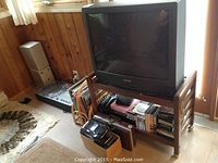 Panasonic 27" CRT TV CT-27D10DB on wooden wheeled stand with shelf containing VHS and DVD players and media, with portable boombox in front and Braun stereo and LP records in background