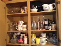 Five cupboard shelves containing assorted dishes and kitchenware, including mugs, glass bowls, pitchers, and utensils.