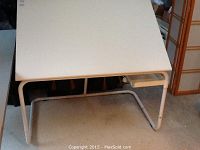 White adjustable art table with rectangular top, swivel supply tray under the top, metal tubular frame, adjustable leg mechanism.
