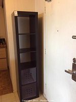 Tall black/brown wood grain shelving unit shown next to a door with four open compartments, two bottom compartments house purple plastic bins.