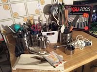 Overview of kitchen utensils and tools placed on a wooden countertop against a tiled wall. Items include cutlery in perforated metal holders, a vegetable slicer, an electric hand mixer, a metal measuring cup, a cream dispenser, scissors, and assorted kitchen utensils.