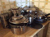 Photo of various sizes of stainless steel pots and pans arranged on a kitchen countertop against a tiled backsplash with circular designs. Includes large, medium, and small pots, frying pan with lid, multi-tier steamer, and flat griddle pan.