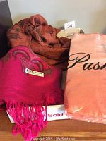 Three pashmina shawls, pink shawl unfolded showing the texture and a wrapped coral shawl to the right with bronze shawl in background.
