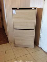 Photo of a light wood IKEA cabinet with two front drop-down compartments for storing shoes or papers, standing on a linoleum floor next to a wall and door.