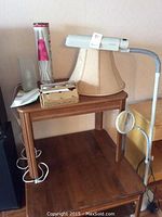View of wooden table with floor-standing ott light with flexible arm and magnifying glass, beige lampshade, boxed lava lamp, light bulbs, and tealight candels on top.