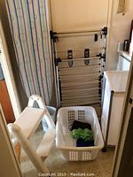 Photo showing ironing board with blue and green circle pattern cover, white plastic laundry basket with clothes inside, white 2-step plastic stool, and metal drying rack against wall.