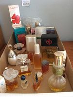 Photo shows approximately 20 assorted perfume and cologne bottles of various brands including Victoria's Secret, Chanel, Bvlgari, Pink Orchid, and others in a cardboard box. Some bottles are new in packaging, others are open.