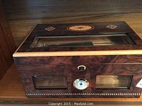 Front view of the wooden humidor showing the lock, hygrometer, glass viewing panels, and decorative inlays on the top.
