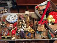 Four trays filled with assorted local and worldly decorative items including ceramics, metal figurines, textiles, and small collectibles.