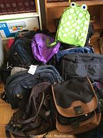 Multiple backpacks, tote bags, computer bags, and duffle bags piled on floor, showing variety of colors including green, purple, black, brown, and pink.