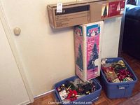 Photo of two boxed artificial Christmas trees standing behind two blue bins filled with Christmas decorations.