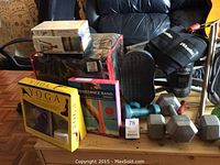 Image showing various yoga and fitness items including yoga tapes, resistance bands, weights, back brace and water bottle arranged on a wooden surface and a chair in the background.