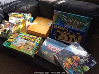 Photo showing collection of box board games Monopoly, Trivial Pursuit, Rummy-O, children's floor puzzle and activity sets displayed on a couch