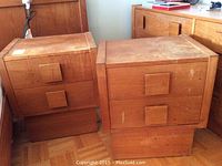 Photo showing two teak bedside tables with visible surface wear, positioned side by side on a wooden parquet floor next to a power outlet and some furniture.