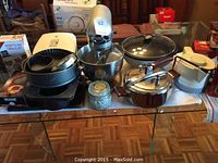 Overview of stand mixer, bakeware stack, grill pan, frying pan, pressure cooker, kettle and Tim Hortons boxes on glass table