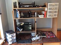 Desk with wooden organizer shelves, desk lamp, Microsoft Arc keyboard, plastic storage drawers, power strips, cables and journals visible on desk surface and shelves.
