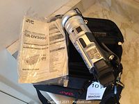 Silver JVC GR-DV3000 digital video camera resting on black JVC carrying case with original instruction manuals in clear plastic sleeve beside it.