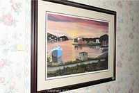 Framed limited edition coastal scene print signed by Ben Gillard hanging on floral wallpaper, showing sunset over harbor with blue boat, red houses, and hills