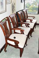 Four wooden dining chairs lined up against a wall near a window. Chairs have curved backs, scroll armrests, and light-patterned upholstered seats.