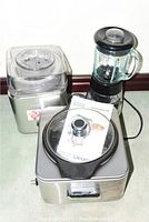 Set of three Cuisinart appliances on floor: ice cream maker with clear lid, blender with glass pitcher and black base, and slow cooker with glass lid and instruction manual on top.