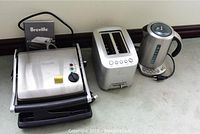 Three appliances side by side: panini grill, toaster, kettle with Breville manual behind grill