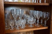 Shelf of assorted crystal stemware including wine glasses, brandy glasses, cordial glasses, and highball glasses with cut glass designs.