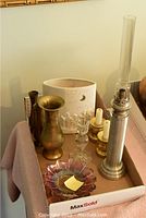 Overview of assorted candle holders, brass vase, and oil lamp arranged in a cardboard tray