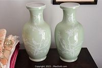 Two light green ceramic vases with white bird and floral branch designs sit side by side on a wooden surface.