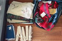 Photo shows a pile of assorted tablecloths including an embroidered cream-colored one, plaid fabric tablecloths inside a black plastic bag, a stack of blue placemats with white edges and floral embroidery, and six rolled cream napkins with decorative napkin rings.