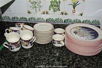 Photo showing sets of large dinner plates, cup saucers, and mugs with Christmas-themed designs and red rims, arranged on a dark granite countertop.