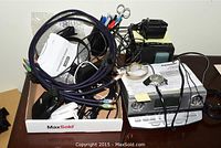 Photo of assorted electronics including cables, headset, watches, and clock radio, all loosely arranged in a box.