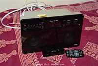 Front and top view of the Tivoli Audio system on patterned fabric showing speaker grills, buttons, and connected cables.