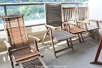 Four outdoor wooden chairs placed in a row on a balcony with views of trees beyond the railing. Chairs show weathering and have slatted backs and seats. One lounge chair with footrest extended is visible.