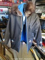Front view of the black nylon parka with hood trimmed in wolf fur, blue lining visible inside, full zipper, and embroidered skiing figure on pockets and sleeves.