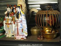 Photo showing two colorful Asian-style ceramic male figures with detailed robes and accessories standing on a shelf beside brass bowls and a decorative black pot with woven details.