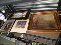 Multiple framed artworks displayed on shelving: includes mountain oil painting, watercolour, print, and tile artwork.