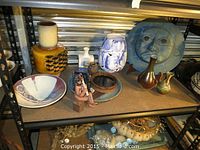 Overall shelf view showing assortment of pottery items including bowls, vases, clay figure, and sun-face plaque.