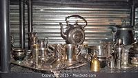 Wide shot of assorted silver plate items arranged on metal shelves including teapot, pitchers, trays, vases, mugs, and small bell.