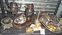Photo of silver plate coffee pot, pitcher, an assortment of decorative silver boxes, a hand mirror, napkin rings, a tray with cutlery, and smaller silver items on industrial storage shelf