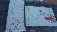 Full view of two framed artworks laid on the ground. One with bird on branch and red flowers, the other floral branch with calligraphy.