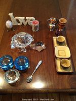 Photo showing full collection on wooden table including stone 'love' sign, vintage egg in brass stand, wooden cup, copper vase, glass turtles, tea light holders, and ceramic tea set.