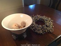 Wide image showing white ceramic bowl on ornate dark metal stand next to grapevine wreath with blue and gold berries, all placed on wooden table.