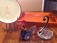 Photo shows a floral hand-painted ceramic plate on a wrought iron stand, a multicolored glass rooster figurine, a vintage glass swan, and a rectangular rubberwood tray in the background.