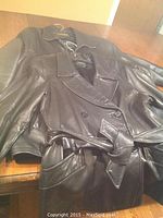 Photo showing two black leather coats on hangers on a wooden table, one trench coat with belt and one leather jacket