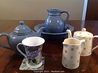 Grey ceramic teapot with lid, pitcher on attached basin, cream and sugar set with pink polka dots, and floral mug with coaster on wooden table.