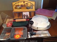 Full view of kitchenware and bakeware set with loaf and cake pans, rolling pin, recipe holder, plastic dome cake cover, pizza pan, spice rack, and kitchen tools on wooden table.