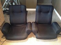 Front view of two black reclining gaming chairs placed side by side on carpeted floor, showcasing armrests and backrests.