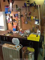 View of pegboard with assorted tools including hammers, saws, clamps, tape measures, utility knives, pipe and tubing cutter, placed against a workbench with various small items and a yellow parts organizer. In front of the bench are a beige box fan, small white desk fan, and stainless steel waste bin.
