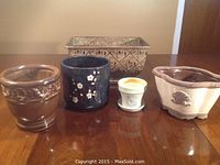 Five planters displayed on a wood surface, showing varied materials, shapes, and colors.